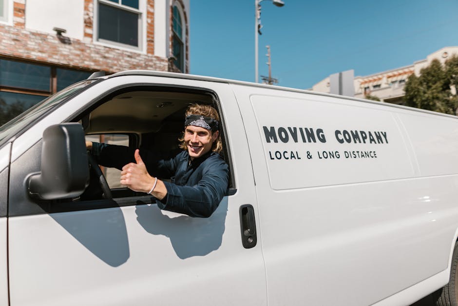A young man with shoulder-length hair, wearing a black bandana and a dark blue shirt, is sitting in the driver's seat of a white moving van branded with 'MOVING COMPANY' and 'LOCAL & LONG DISTANCE' text on the side. He is smiling and giving a thumbs-up gesture through the open driver’s side window. The van is parked on a residential street with multi-storey brick buildings, a clear blue sky, and a utility pole visible in the background. Inside the van, visible through the open door, are cardboard boxes, some wrapped in plastic or fabric, along with furniture such as a wooden chair and wrapped cushions, indicating an ongoing home relocation process. The scene suggests a professional removals service preparing for or engaged in furniture transport and packing during a house move, with equipment like trolleys or straps possibly nearby out of view. Finchley Removals specializes in local and long-distance moving logistics, as exemplified in this loading process on a street with tight parking or narrow streets, adhering to best practices for house removals and furniture transport.