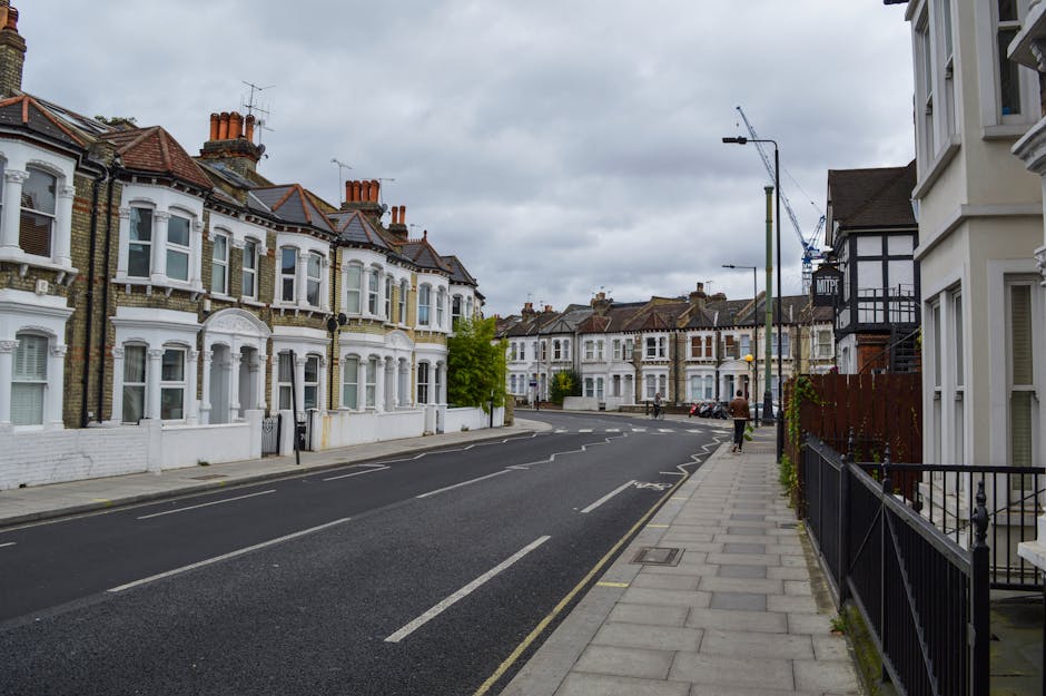 A street view of residential houses on a cloudy day, with a sidewalk on the right side featuring a black metal railing and a row of white terraced homes with bay windows and brick detailing. The road has dashed white lines and no parked vehicles, while a few pedestrians are visible walking along the pavement. In the background, construction cranes are seen above additional row houses, indicating ongoing development nearby. The scene captures a typical urban neighbourhood environment, suitable for house removals or home relocation activities. Finchley Removals operates in this area, handling furniture transport, packing, and loading processes for local house moves.