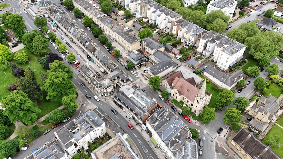 An aerial view of North Finchley High Road showing a busy street with lined parked cars on both sides, surrounded by rows of residential buildings and small commercial properties. The pavement is populated with pedestrians, and there are multiple delivery vans and service vehicles parked along the street. Trees with lush green foliage line the sidewalks and fill nearby open spaces, providing a contrast to the urban environment. In the foreground, a building with a shopfront and a small parking area is visible, while further along the road, a mix of terraced and semi-detached houses can be seen. A historic church with a pointed steeple and stained glass windows is situated near the residential area, amidst the greenery and surrounding dwellings. The overall scene captures a typical day of local activity and movement on North Finchley High Road, reflecting the busy logistics involved in house removals and furniture transport services, as coordinated by Finchley Removals.