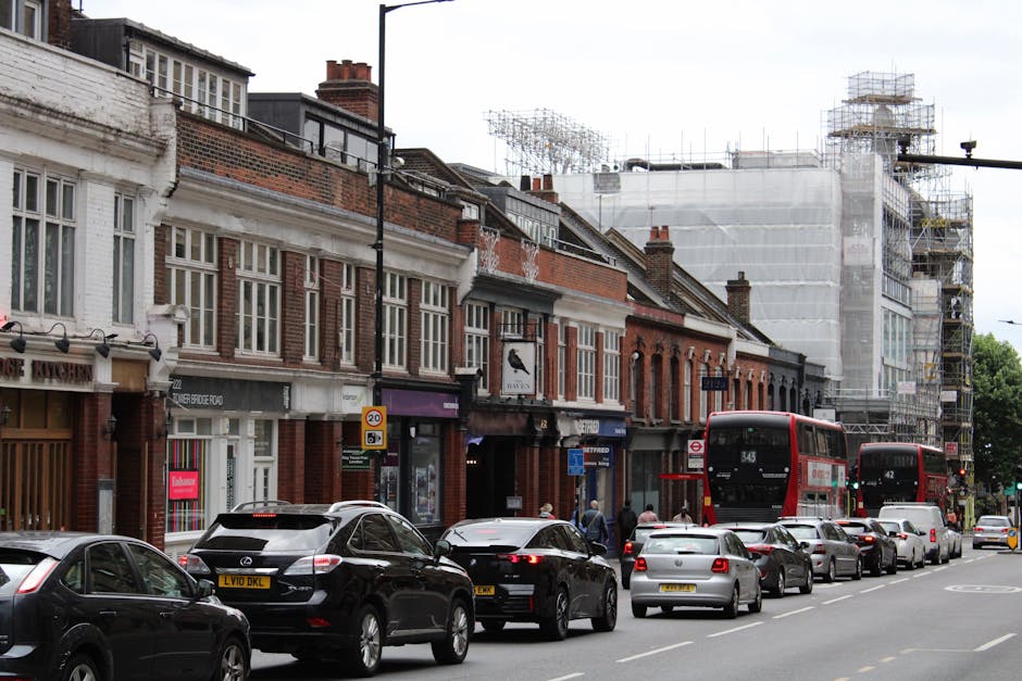 Street view of North Finchley High Road showing a row of brick and white-painted commercial buildings with large windows, some with signage for local shops and cafes. In front of the buildings, there is a line of parked cars, including a black SUV, a grey sedan, and other small vehicles. Two red double-decker buses are stopped along the kerb, with one bus displaying the route number 343. To the right, a construction site is visible, covered in scaffolding and white protective sheeting, indicating ongoing building work. Pedestrians are walking along the sidewalk, some near the shop entrances. The scene is captured during daylight with overcast weather, and the environment suggests an active urban area typical of a busy town or city centre. Finchley Removals' loading process may involve navigating streets like this for home relocation, furniture transport, and packing and moving services around North Finchley High Road.