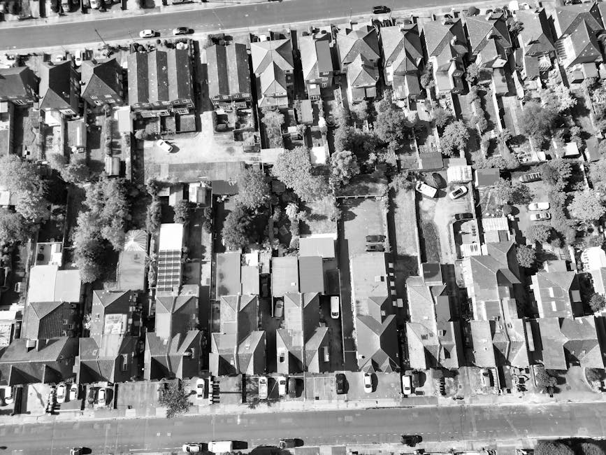 An aerial view of North Finchley High Road showing a busy street with lined parked cars on both sides, surrounded by rows of residential buildings and small commercial properties. The pavement is populated with pedestrians, and there are multiple delivery vans and service vehicles parked along the street. Trees with lush green foliage line the sidewalks and fill nearby open spaces, providing a contrast to the urban environment. In the foreground, a building with a shopfront and a small parking area is visible, while further along the road, a mix of terraced and semi-detached houses can be seen. A historic church with a pointed steeple and stained glass windows is situated near the residential area, amidst the greenery and surrounding dwellings. The overall scene captures a typical day of local activity and movement on North Finchley High Road, reflecting the busy logistics involved in house removals and furniture transport services, as coordinated by Finchley Removals.